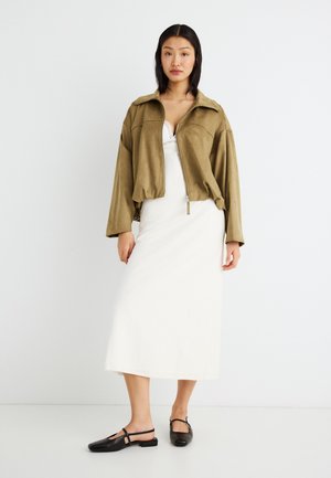 Kvinde iført en lysebrun lynlåssweater over en hvid midi-kjole og sorte slingback-flats, stående mod en ensartet hvid baggrund.