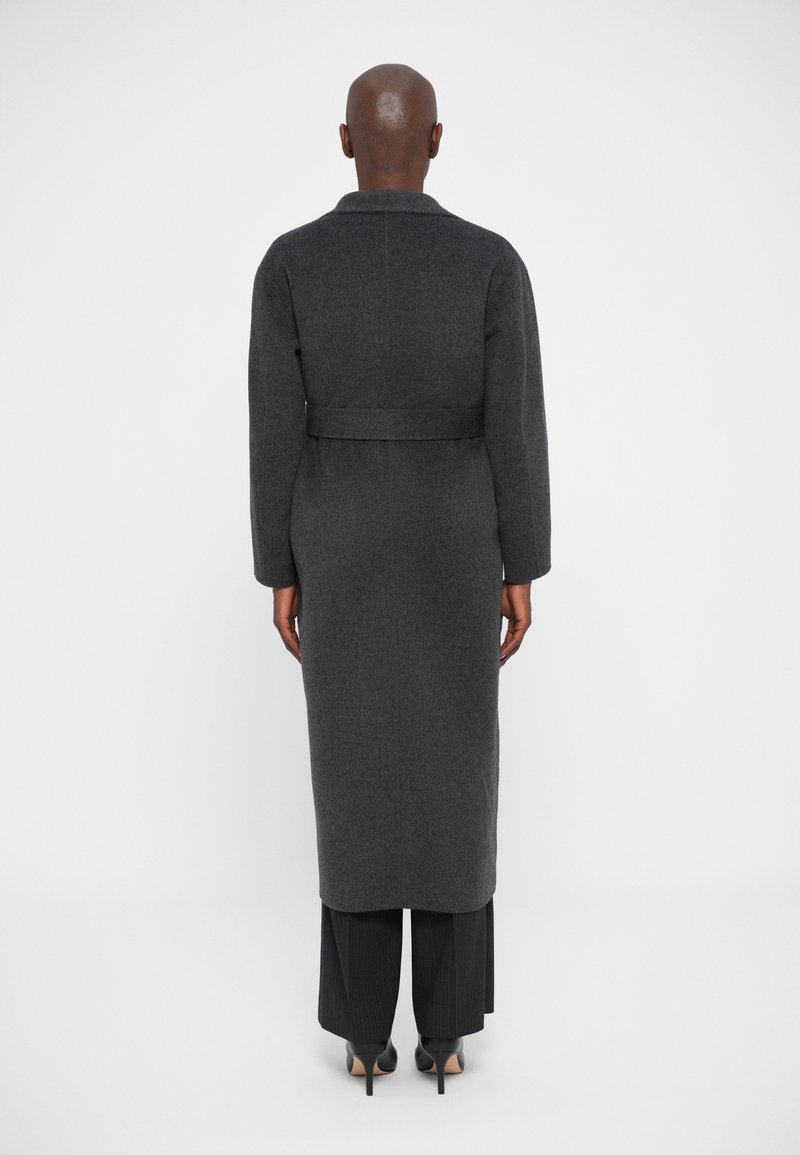 Person wearing a long dark gray belted coat, black pinstripe pants, and black high-heeled shoes, standing facing away on a white background.