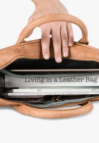 Hand holding open tan leather bag showing a book titled "Living in a Leather Bag," magazines, and a wallet inside.