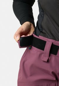 Lila Gürteltasche mit schwarzer Klettverschluss, befestigt an strapazierfähigen, leichten Hosen. Die Textur ist glatt; zeichnet sich durch ein kleines blaues und gelbes Etikett aus.