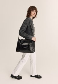 Sac à main en cuir noir avec deux poignées et une bandoulière amovible, comportant une poche zippée et un détail de logo, présenté avec un pull en maille gris et un pantalon blanc.