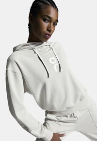 Hoodie beige fabriqué en tissu doux avec une poche avant, présentant un grand logo blanc sur la poitrine et des cordons de serrage, associé à un pantalon assorti.