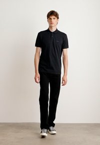 Μαύρο πόλο μπλουζάκι με κοντά μανίκια και γιακά, με διακριτό λογότυπο, σε συνδυασμό με μαύρο παντελόνι και sneakers σε μαύρο και άσπρο χρώμα.