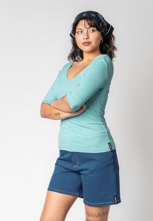 Helles türkisfarbenes, geripptes Oberteil mit drei Viertelärmeln, kombiniert mit dunkelblauen Shorts. Das Oberteil hat einen runden Ausschnitt und eine leichte Textur.