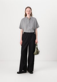 Zwarte wijde broek, gecombineerd met een gestreepte korte mouwen overhemd met knopen en een gestructureerde olijfgroene handtas. Platte zwarte schoenen.