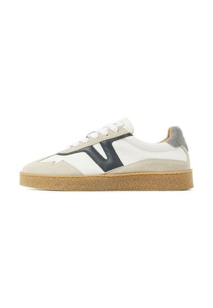 Witte lage sneaker met beige suède accenten, zwart zijlogo, witte veters, grijze hielpatch en een lichtbruine geribbelde rubberen zool.