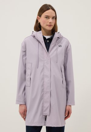 Jonge vrouw die een lichtpaarse regenjas met capuchon draagt over donkere kleding, staand tegen een eenvoudige lichte achtergrond.