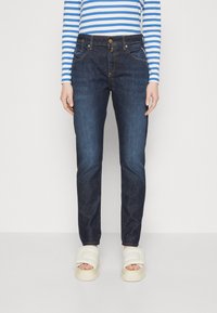 Jeans in denim blu scuro con vestibilità slim, dotati di tasche frontali e chiusura a bottone visibile. Indossati con sandali con platform chiari.