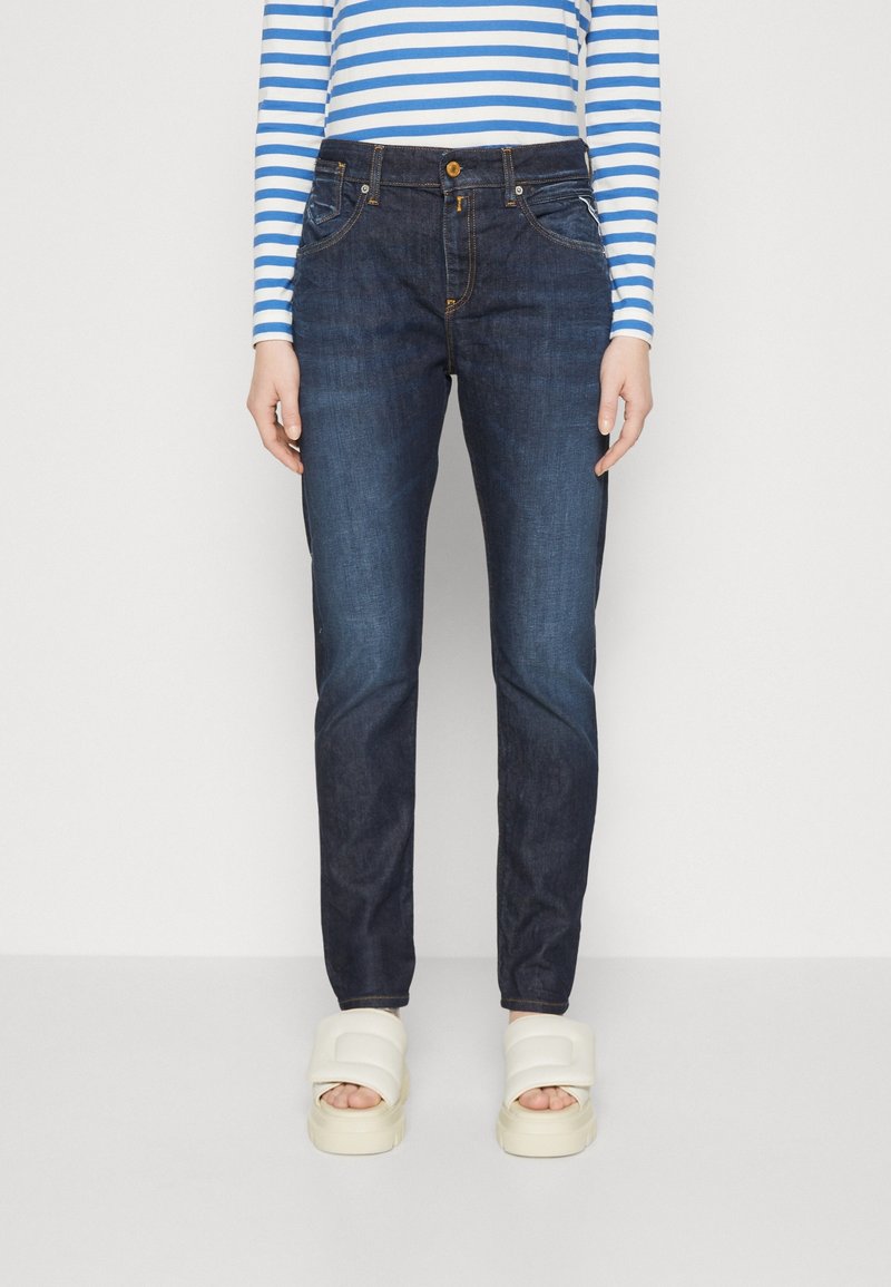 Jeans in denim blu scuro con vestibilità slim, dotati di tasche frontali e chiusura a bottone visibile. Indossati con sandali con platform chiari.