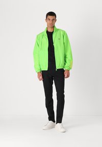 Casaco verde neon com fecho de correr, punhos elásticos e uma textura leve, combinado com calças pretas e ténis brancos.
