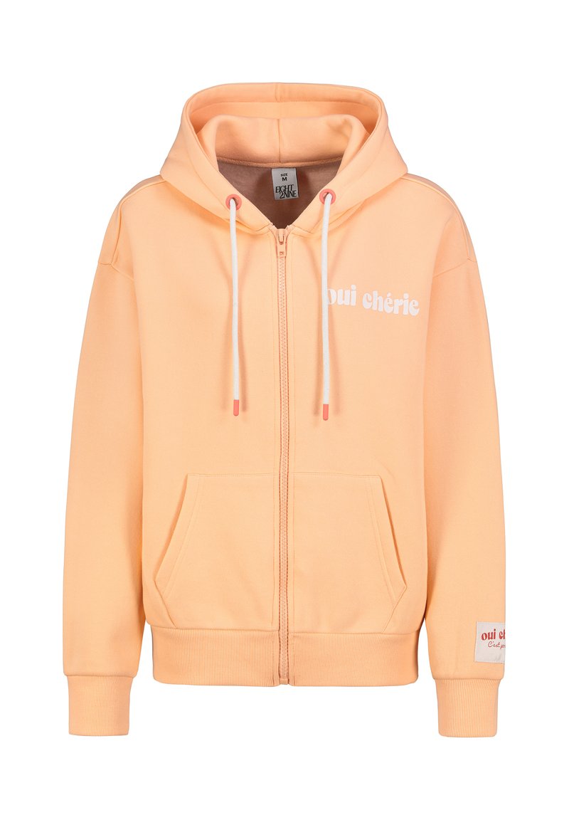 Pfirsichfarbener Kapuzenpullover mit Frontreißverschluss, Seitentaschen, weißem Schriftzug "oui chérie" und kontrastierenden Kordeln mit abgedeckten Enden. Weiches Material.