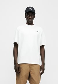 Άσπρο κοντομάνικο μπλουζάκι με μικρό μαύρο λογότυπο στο στήθος, σε συνδυασμό με παντελόνι ταμπά και σκούρο μπλε καπέλο με λευκό λογότυπο.