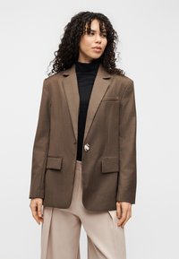 Blazer marron sur mesure avec un seul bouton, deux poches avant et des épaules structurées. Porté sur un col roulé noir. Texture de tissu lisse.