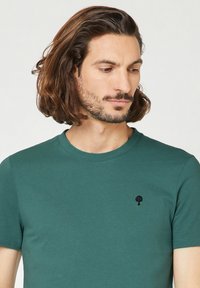 T-shirt in cotone verde con maniche corte, colletto rotondo e un piccolo logo nero a forma di albero sul petto. Texture liscia, design aderente.