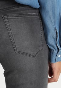 Jeans de mezclilla gris oscuro con un detalle en el bolsillo trasero, combinados con una camisa de cuello azul claro con puños abotonados. Textura suave, diseño ajustado.