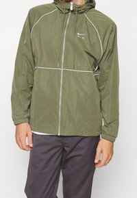 Olivegrön lättviktsjacka med huva, dragkedja framtill och elastiska ärmslut. Har vita accenter och en reflekterande logotyp på bröstet.