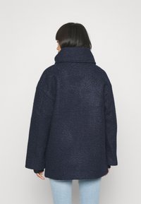 Marinblå, överdimensionerad kappa med hög krage och textil med struktur. Har sänkta axlar och långa ärmar, designad för komfort och värme.