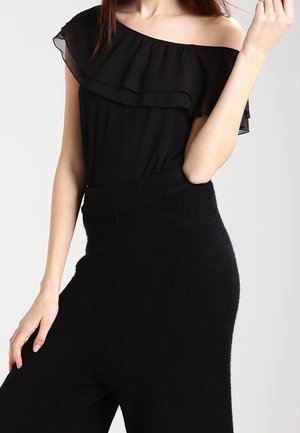 Vrouw draagt een zwarte off-shoulder top met ruches en hoog getailleerde zwarte geaderde broek tegen een effen achtergrond.