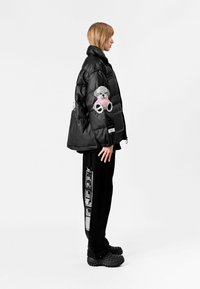 Chaqueta acolchada negra con un acabado texturizado, que cuenta con un parche de un oso de peluche rosa en la manga. Combinada con pantalones negros y zapatos negros de suela gruesa.