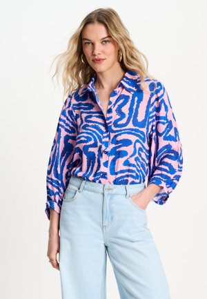 Vrouw draagt een roze en blauw abstract patroon blouse met knopen en lichtblauwe high-waist jeans, staand met één hand in de zak.