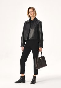 Giacca cropped in pelle nera con chiusure a scatto, abbinata a pantaloni neri aderenti e stivaletti alla caviglia. Borsa in pelle marrone con una texture e hardware dorato.
