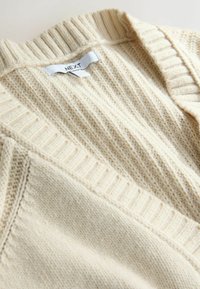 Kreemi värvi kootud kardigan, millel on ribakujundus, v-kaelus ja tekstuuriga kangas. Silt on nähtav taga.