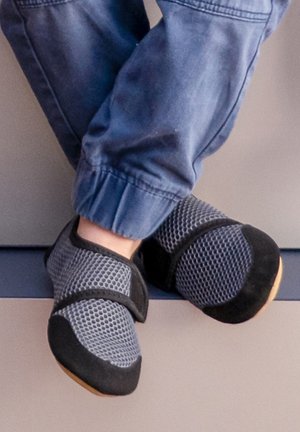 Graue Mesh-Hausschuhe mit schwarzen Akzenten, ausgestattet mit einem Klettverschlussriemen und einer flexiblen Sohle, kombiniert mit blauen, umgeschlagenen Hosen.