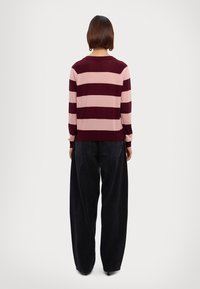 Pull rayé bordeaux et rose avec manches longues, col rond et poignets côtelés. Associé à un pantalon noir à coupe ample.