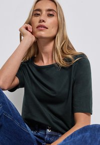 Tmavě zelené tričko s krátkým rukávem vyrobené z jemné látky, s kulatým výstřihem. Model pózuje nenuceně v džínových kalhotách.