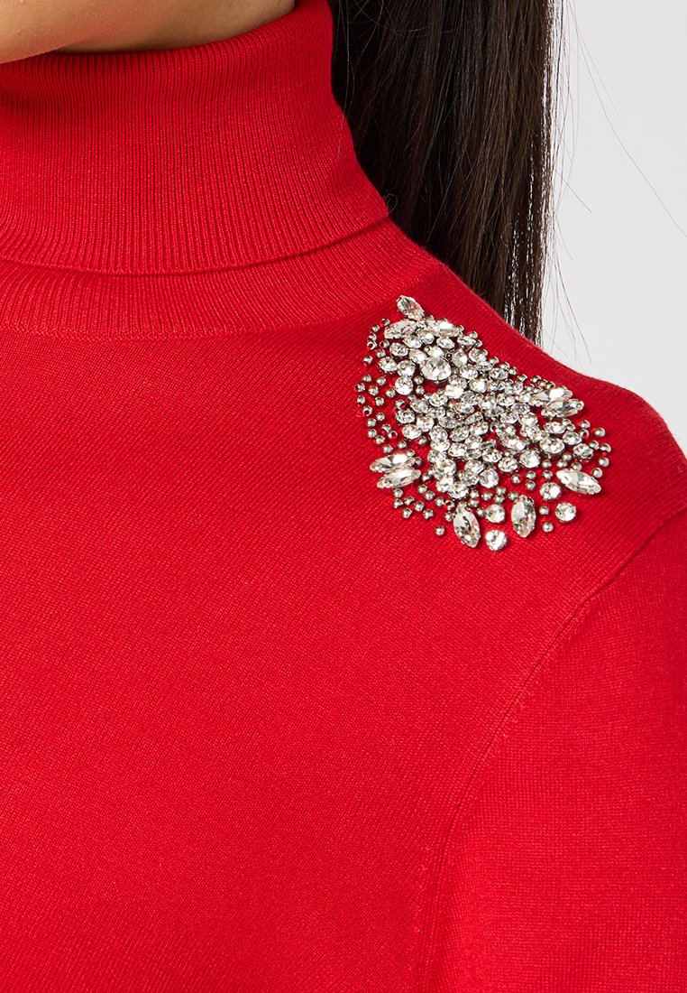 Maglione a collo alto rosso realizzato a maglia con un'applicazione di strass sulla spalla sinistra, che aggiunge texture e brillantezza al tessuto.