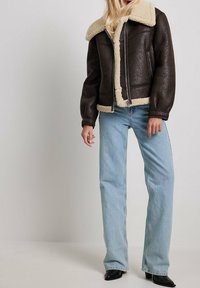 Kvinde iført mørkebrun lammeskindsjakke, lyseblå jeans med lige ben og sorte støvler med spids næse, stående mod en ensfarvet baggrund.