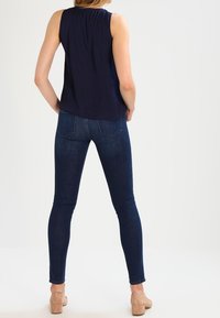 Granatowa bezrękawnik z marszczonym dekoltem w połączeniu z ciemnoniebieskimi rurkami i jasnobrązowymi sandałami na kostkę, widok z tyłu.