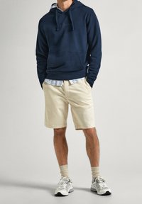 Man som har på sig en marinblå huvtröja, beige shorts, en randig skjorta under, beiga strumpor och vita sneakers, står med händerna i fickorna.