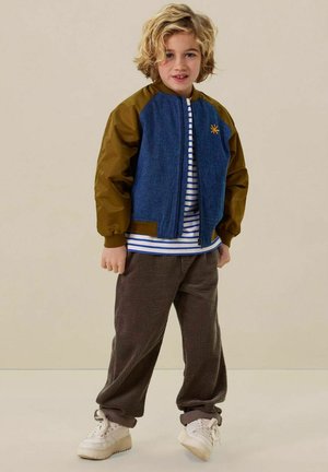 Denimjack met bruine mouwen, ritssluiting aan de voorkant en zon-embleem. Gepaard met gestreept shirt, bruine corduroy-broek en witte sneakers.