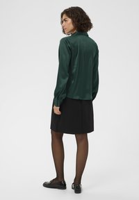 Green satin blouse with a collar, long sleeves, and button cuffs, paired with a black skirt and black shoes. Smooth texture and simple design.