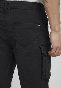 Pantalon en denim noir à coupe droite, avec une poche arrière ornée d'un motif chevron cousu et une poche cargo latérale.