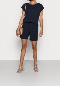 Osoba w czarnej bluzce z krótkim rękawem i pasujących czarnych szortach, w jasnoniebieskich sandałach na obcasie i niosąca beżową torebkę na ramię.