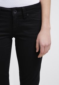 Černé džíny z denimu s přiléhavým střihem. Mají jedno zapínání na knoflík a přední kapsy. Hladký povrch a minimální švy.