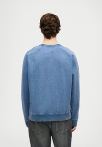 Hombre con cabello rizado castaño, vistiendo una sudadera azul y jeans negros desgastados, de espaldas, contra un fondo claro y liso.