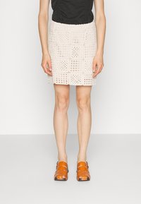 Mini spódniczka na szydełku w jasnobeżowym kolorze z kwadratowymi wzorami i falowanym brzegiem. Połączona z pomarańczowymi sandałami na paski.