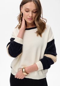 Roomkleurige pullover met marineblauwe en beige gestructureerde mouwen. Voorzien van geribbelde manchetten en zoom, met een zachte afwerking. Eenvoudig en modern ontwerp.