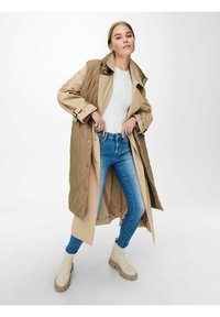 Beige og olivenfarget quiltet lang jakke over en hvit skjorte, kombinert med blå smale jeans og kremfargede ankelstøvler. Glatt stoff, avslappet passform.
