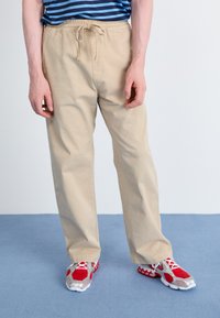Beige cotton pants with an elastic waistband and drawstring, featuring a relaxed fit; paired with red and silver athletic shoes.