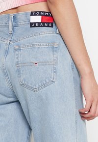 Los jeans de mezclilla de color azul claro cuentan con un bolsillo trasero que lleva una pequeña etiqueta con el logo. La cinturilla muestra una etiqueta con "TOMMY JEANS" en colores contrastantes.