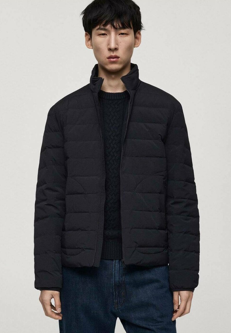 Veste matelassée noire avec un col haut, fermeture éclair et deux poches avant, associée à un pull en tricot sombre et un jean bleu.