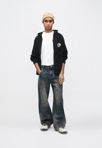 Μαύρη πλεκτή ζακέτα με φερμουάρ, λευκό t-shirt, φαρδιά ξεθωριασμένα τζιν παντελόνια και κρεμ παπούτσια. Μπεζ σκουφί με διακριτικό μοτίβο.