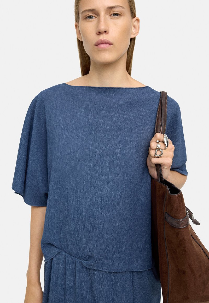 Niebieska teksturowana bluzka z dekoltem łódkowym i luźnymi rękawami, zestawiona z brązową torebką z zamszu z skórzanymi akcentami.