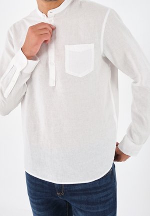 Homme en train de fermer le dernier bouton d'une chemise en lin blanche à manches longues avec une poche sur la poitrine, portant un jean bleu, sur un fond blanc uni.
