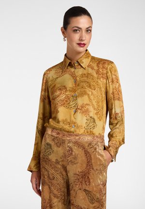 Frau trägt ein passendes goldenes Paisley-Muster langärmeliges Hemd und Hose, mit einer Hand in der Tasche vor weißem Hintergrund.