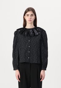 Svart bluse med en paljettbesatt volangkrage, teksturert stoff, hvite prikker og knappeåpninger, med lange puffede ermer.